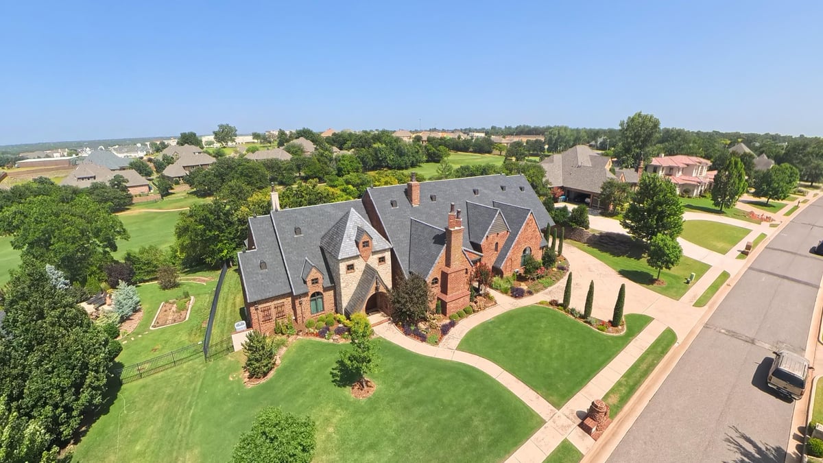 Aerial view of a large brick mansion with gray roof surrounded by manicured lawns and trees in a suburban residential area