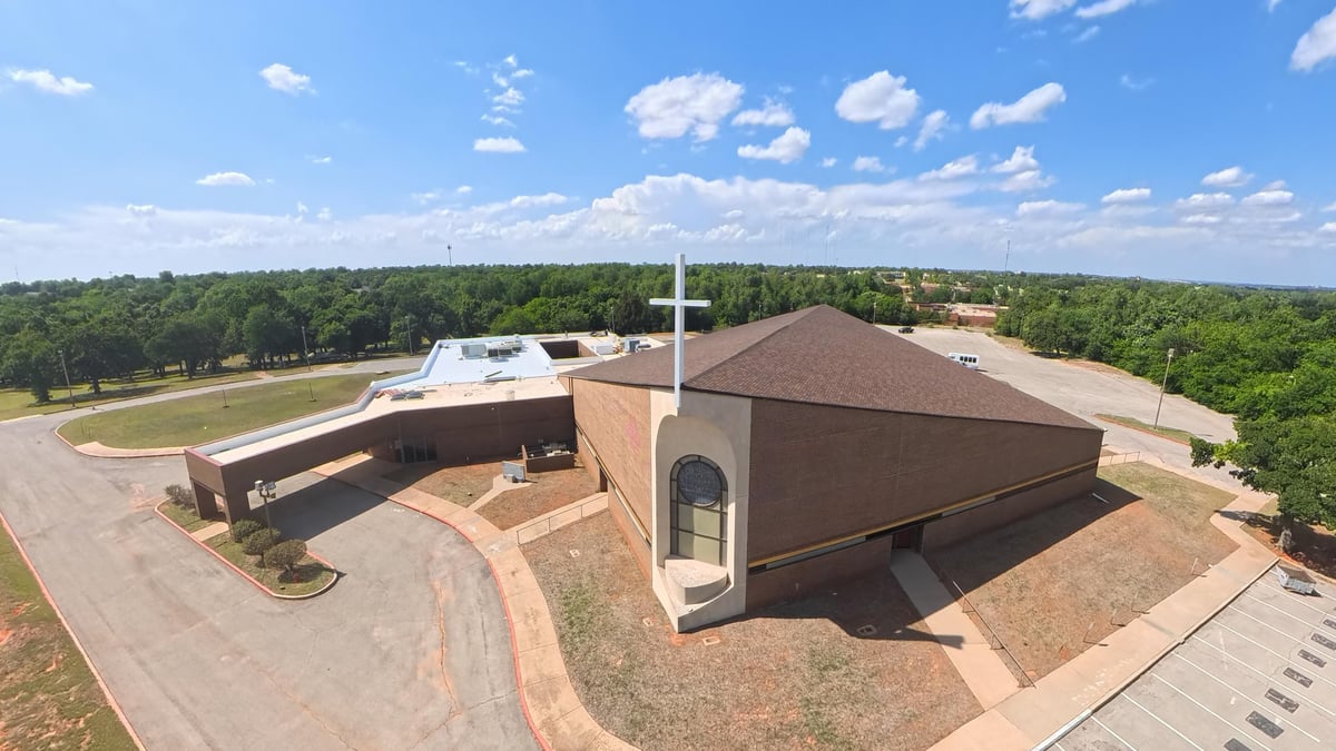 Aerial view of a modern brown brick building with a curved design and white cross, surrounded by landscaping and green forest