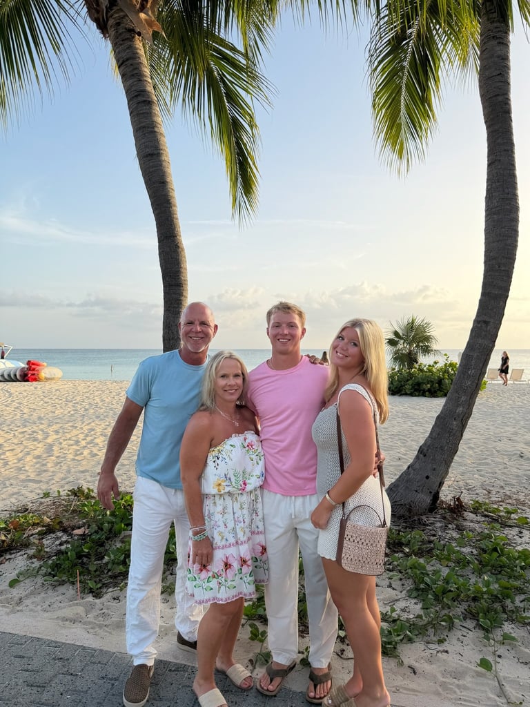 Four people posing together on a tropical beach with palm trees, ocean, and sandy shore in the background