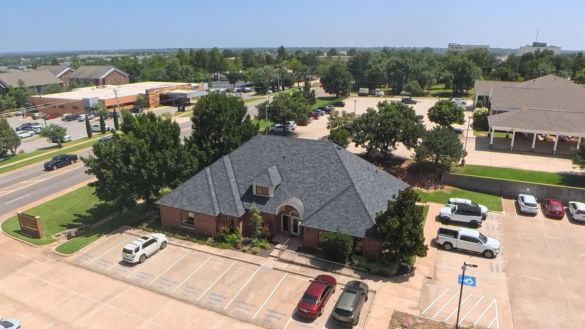 Aerial view of a brick building with dark roof in a suburban area surrounded by parking lots, trees, and residential homes